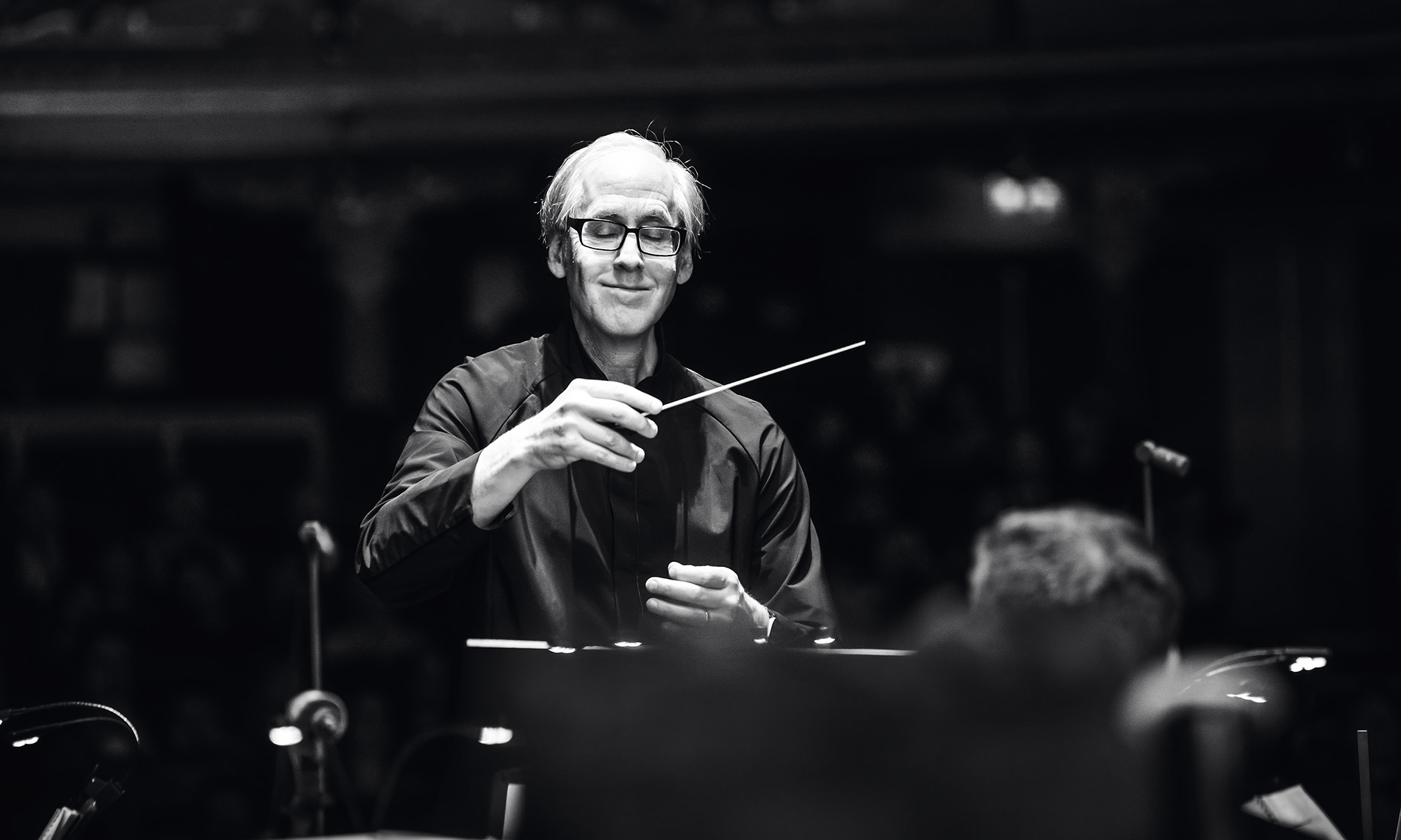 Composer Jeff Beal smiles while conducting, holding a baton in a black-and-white portrait.