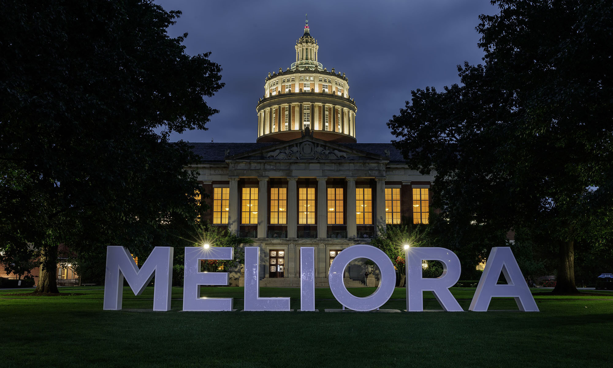 MELIORA letters and Rush Rhees Library lit up during twilight on the Eastman Quad.