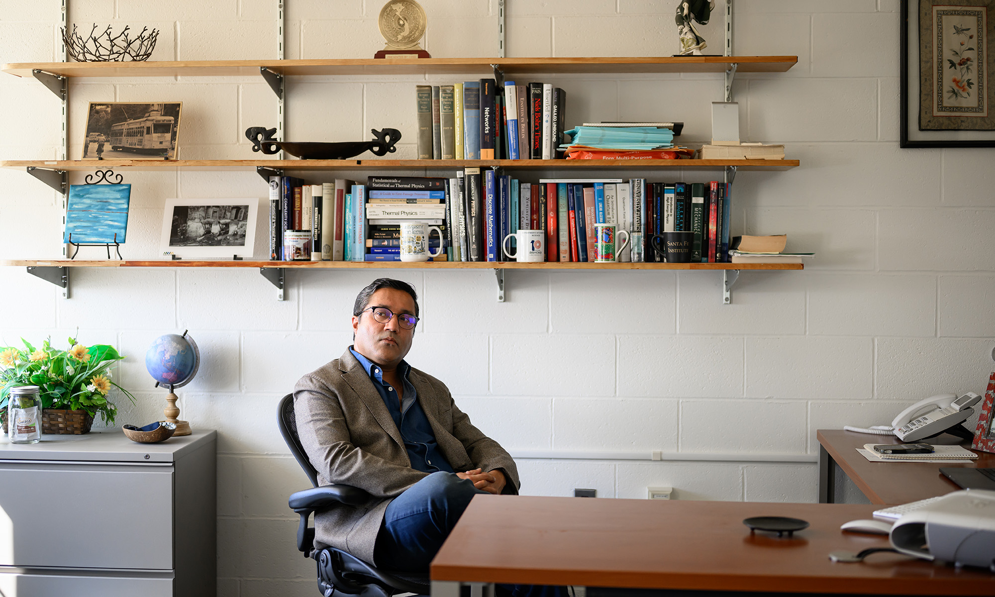 Gourab Ghoshal sits at his desk in his office with a bookshelf full of books and objects behind him.
