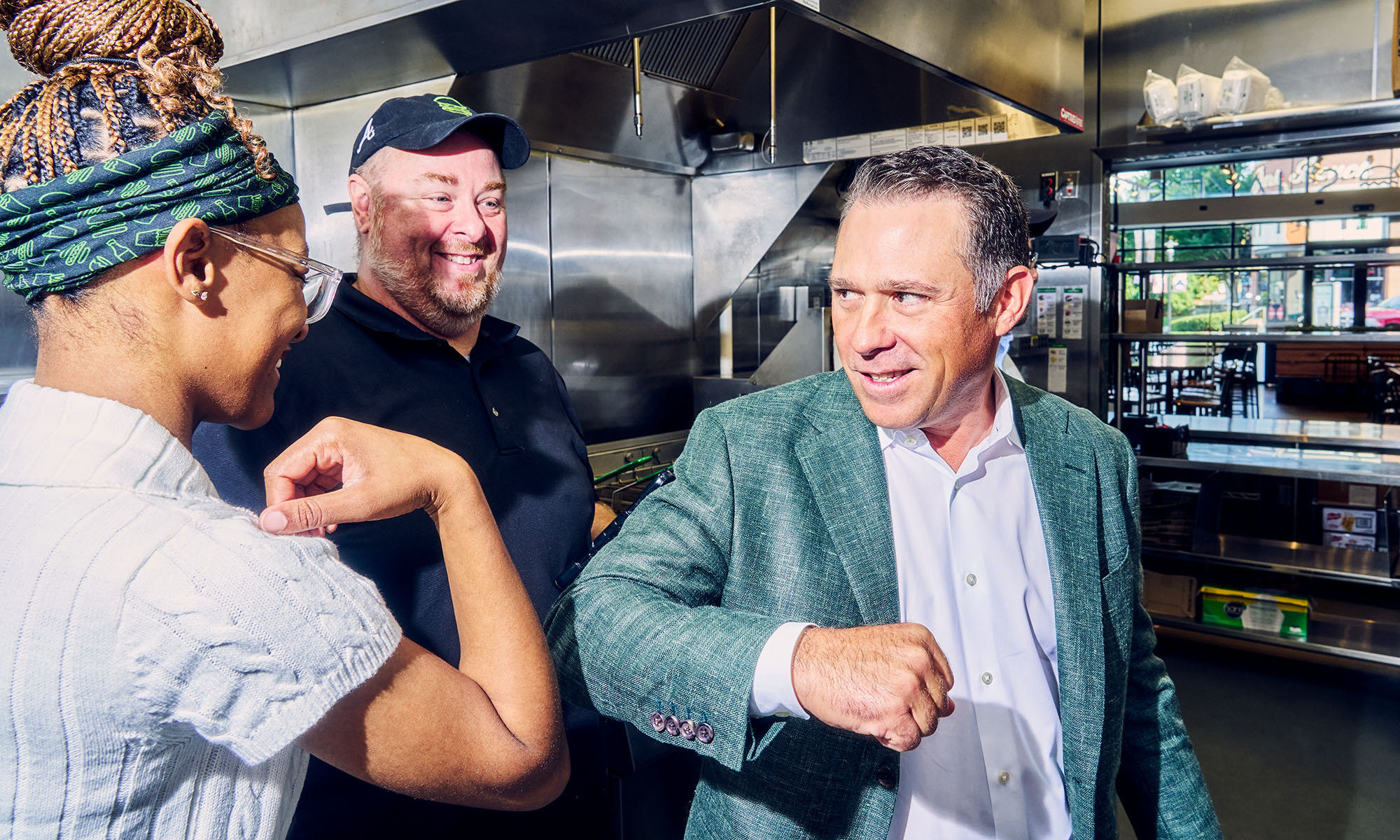 Shake Shack CEO Rob Lynch, wearing a green blazer, exchanges an elbow bump with an employee in the restaurant kitchen as another employee smiles.