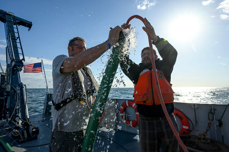 Two crew members lift and position a large hose connected to the ship's water-sampling equipment.