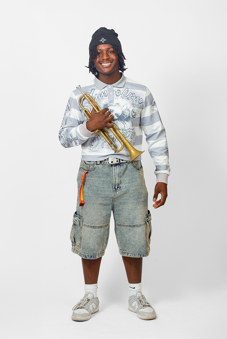 Nate France in white shoes, light-washed jean shorts, and a gray and white horizontal-striped rugby-style shirt holds a trumpet.