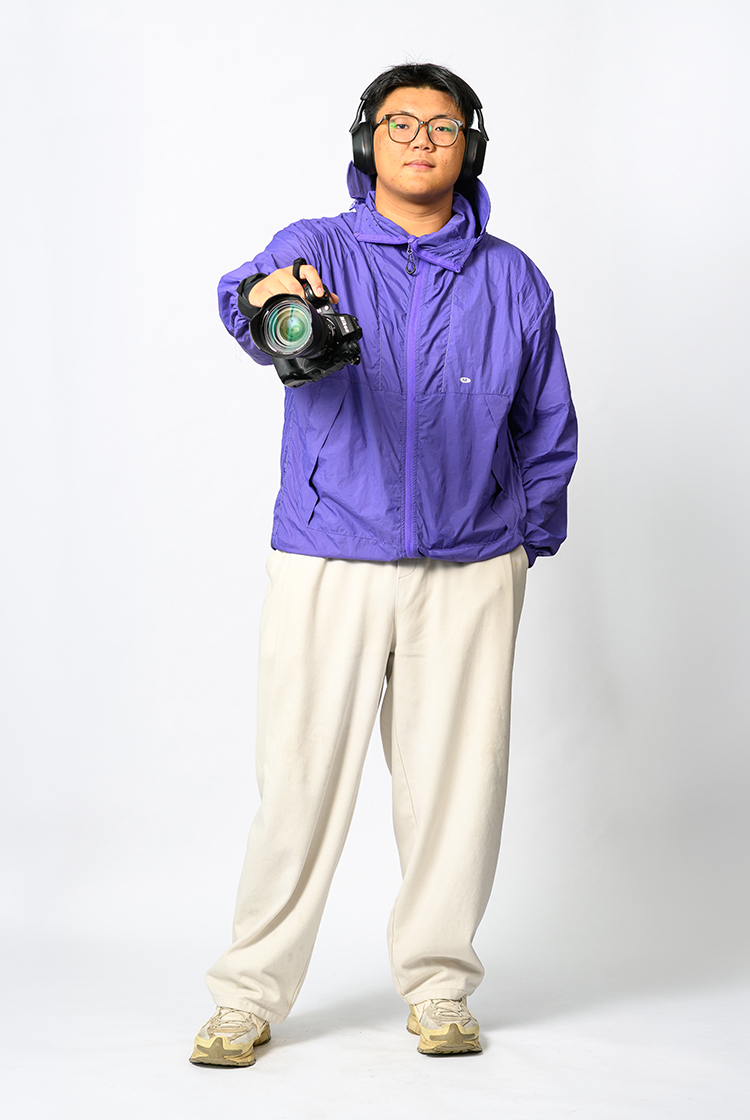 Dede Xu in a bright purple windbreaker, white sweat pants, white sneakers, with headphones on as he holds a camera to the camera.