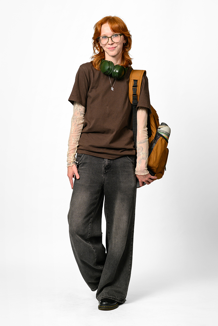Karis Kelly sporting a see-through long-sleeve shirt with a brown t-shirt over it, black jeans, green headphones, and a brown backpack.