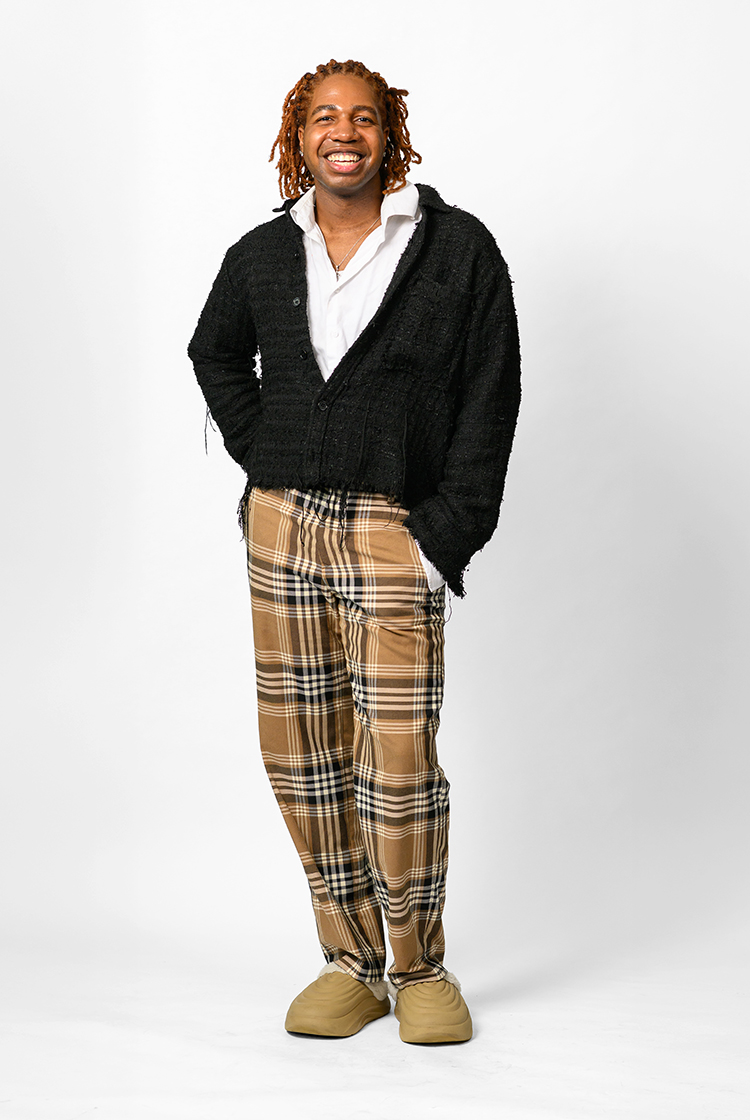 Kevin Johnson in a white button down shirt open at the first three buttons, an oversized black cardigan, brown plaid pants, and taupe, clog-like shoes.