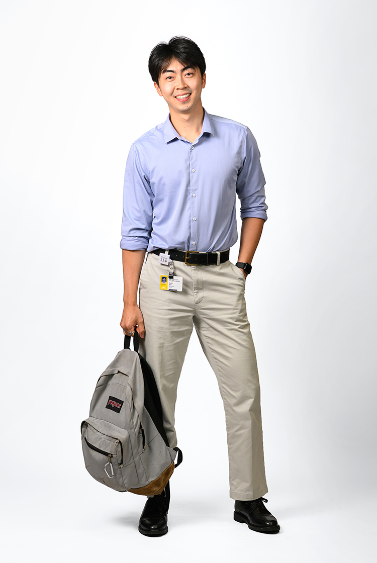 David Zhu in a blue, long-sleeve, button-down shirt, khakis with black belt and shoes, holds his gray Jansport backpack.