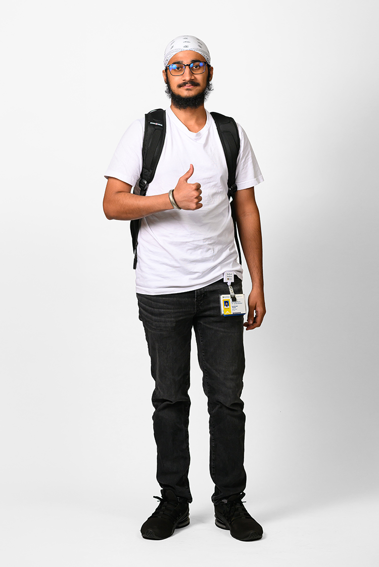 Sukhman Thind in a white v-neck shirt, black pants with his URochester Medicine ID clipped on it, black shoes, and a black backpack gives a thumbs up.