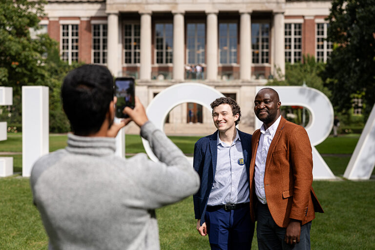 Two Meliora Weekend attendees pose for a photo while a third captures a cell phone photo of them in front of the MELIORA letters.