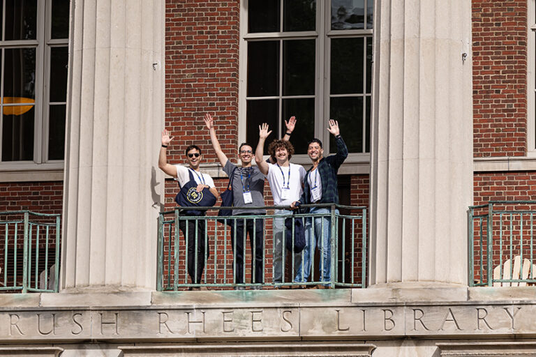 Four young male Meliora Weekend attendees stand on the Rush Rhees Libary balcony and wave at the camera.