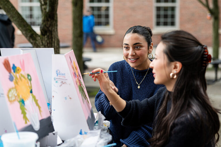 Two women by an easel paint a painting of flowers in a vase.
