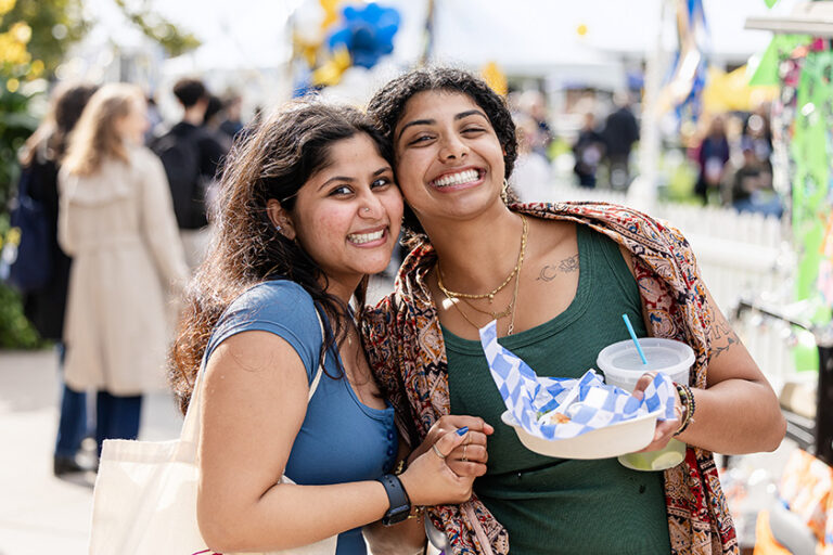 Two women beam at the camera while holding their food in the Meliora Village.