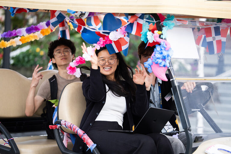 Three Meliora Weekend attendees wave while driving by in a decorated golf cart.
