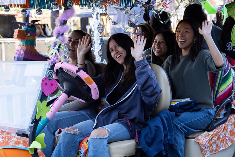 Four young women in a decked out golf cart wave at the people cheering them on.