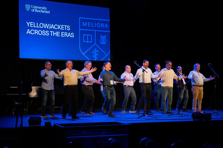 Members of the YellowJackets a cappella group from decades ago perform on stage with a screen behind then that reads 