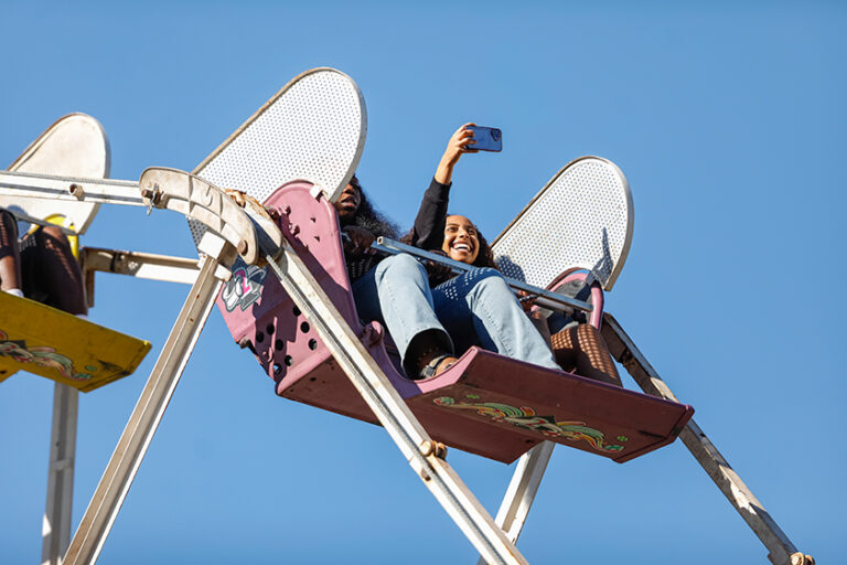 Two people in a Ferris wheel gondola take a selfie against a bright blue sky.
