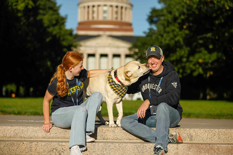 A young woman, a yellow lab, and a middle aged man decked out in URochester gear sit on the Eastman Quadrangle steps with Rush Rhees Library in the background.