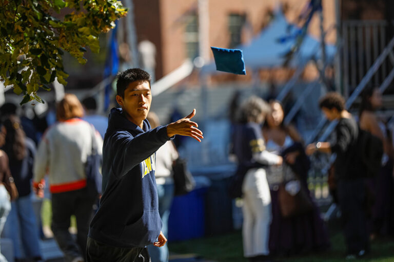 Meliora Weekend attendees throws a cornhole beanbag surrounded by fellow attendees in the Meliora Village area.