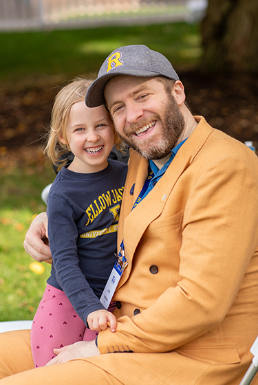 A URochester alumnus in a yellow sport jacket smiles while holding a child in a Yellowjacket shirt.