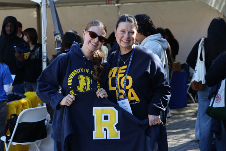 Two University of Rochester undergraduates in blue-and-yellow gear hold up a blue sweatshirt with a giant yellow 