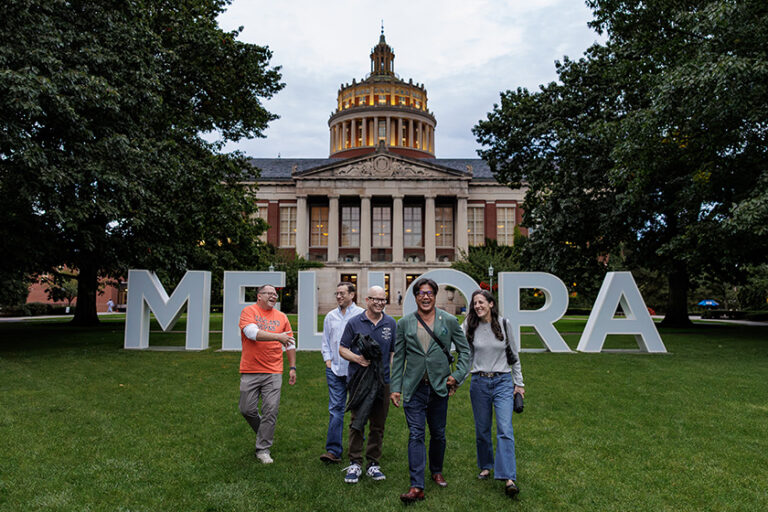 A group of Meliora Weekend 2026 attendees walk toward the camera from the giant MELIORA letters as the sun sets.