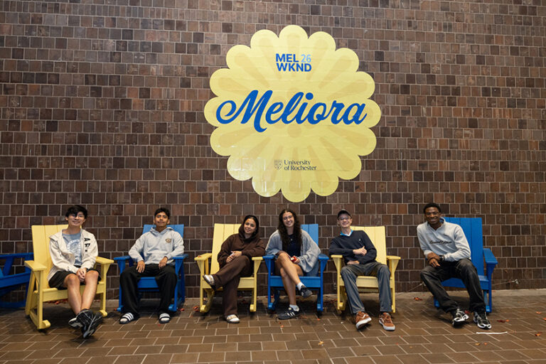 People sitting in blue and yellow Adirondack chairs in front of a giant yellow dandelion decal that reads Meliora in blue font.