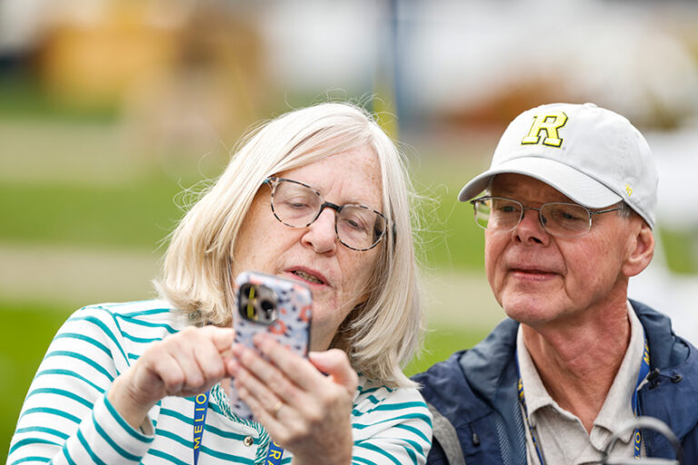 An elderly woman shows off something on her phone to the elderly man sitting next to her and sporting a University of Rochester cap.