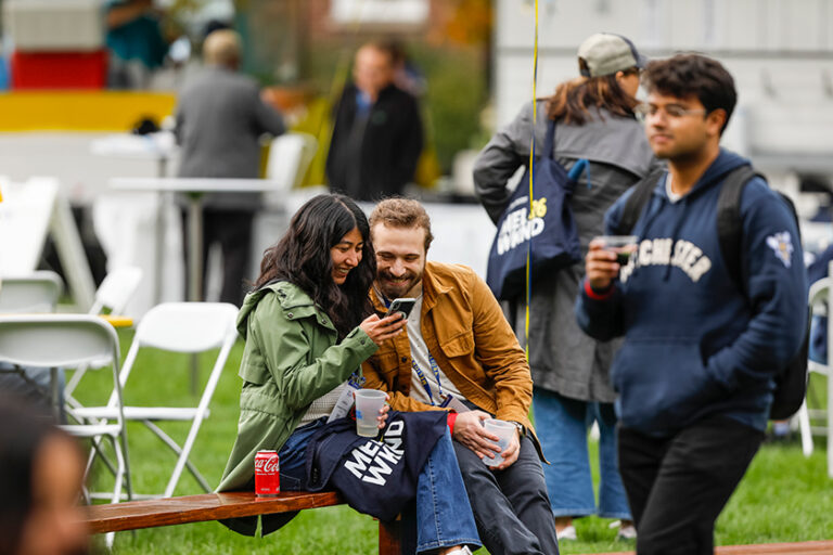 A woman shows something on her phone to a man sitting next to her while Meliora Weekend attendees walk nearby.
