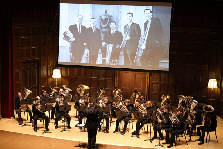 Tuba and other horn instruments players seen from above with a screen behind them showing a black-and-white archival photo of five men in suits with their horn instruments.