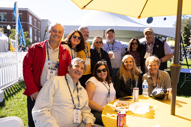 Nearly a dozen attendees sit together and smile for a group photo at a table with a big yellow umbrella overhead.