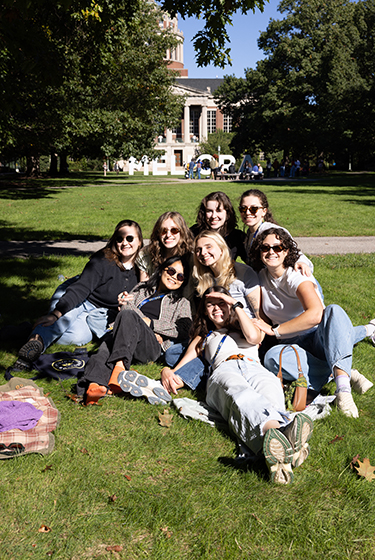 A big group of young women awash in sunshine and huddled together while smiling on the Eastman Quad.