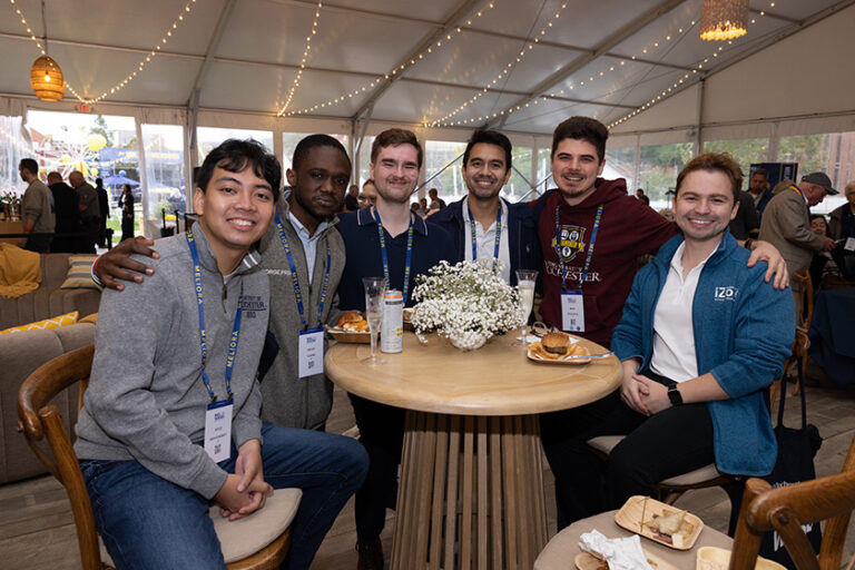 Six alumni gathered around a round table under the Meliora Village tent smile at the camera.