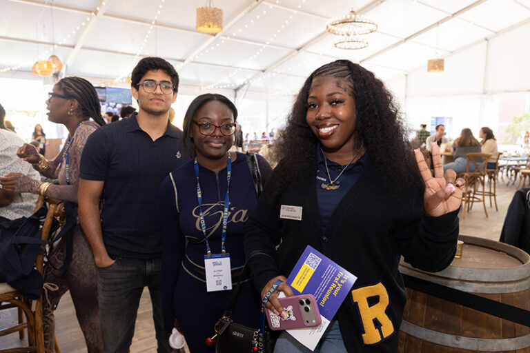 Two women and a man in Rochester blue gear smile at the camera with one making a peace sign gesture.
