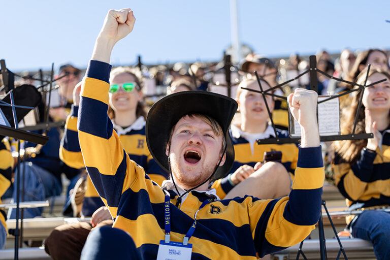 Close-up of a fan wearing a blue and yellow striped rugby shirt cheering boisterously while watching the URochester football game.