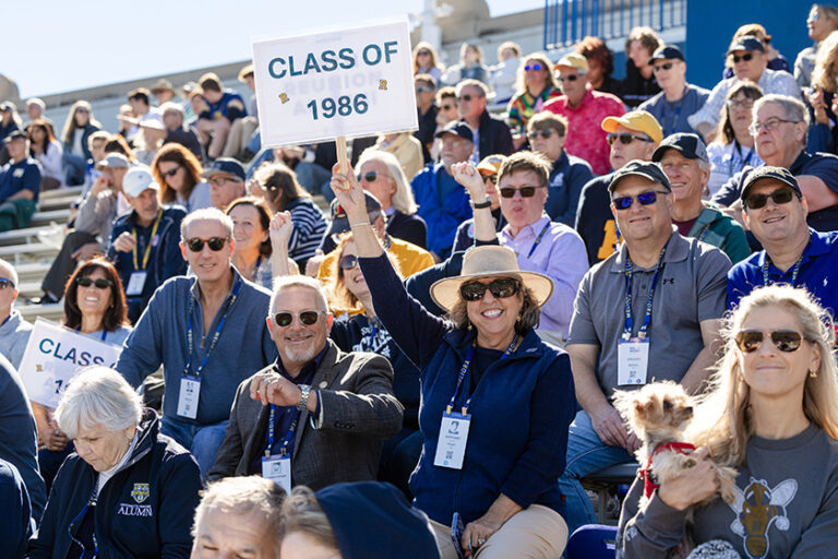 Football stadium seats filled with URochester alumni and supporters, including one woman holding up a sign that says 