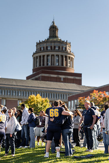 A URochester football player in uniform seen from behind getting hugged while tailgaters celebrate a Yellowjacket victory and Rush Rhees Library towers overhead.