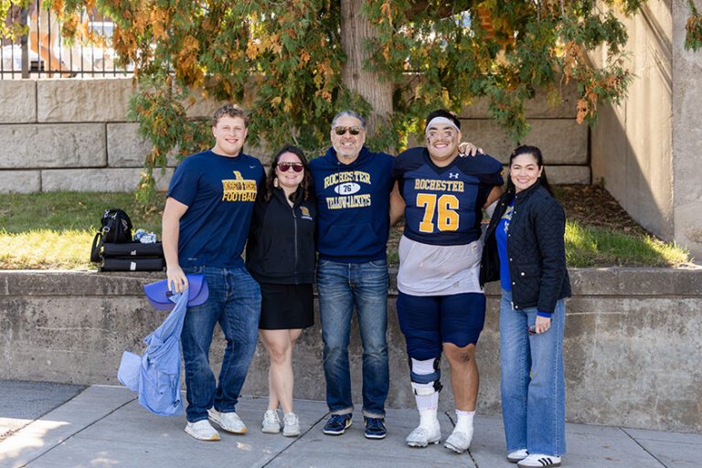 Five Meliora Weekend attendees decked out in URochester gear smile for a photo with their arms around each other.