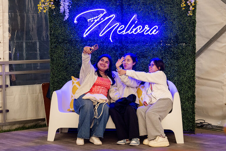Three young women take a selfie on a couch below a neon light that reads 