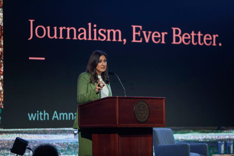 Amna Nawaz at a podium on stage in front of a large screen that reads 