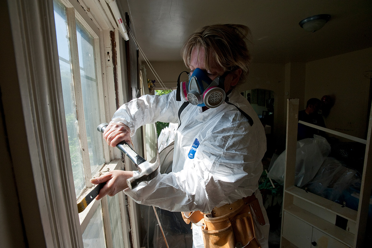 A worker wearing a protective suit and respirator uses a hammer and pry bar to remove an old window frame during lead-paint abatement in a home.