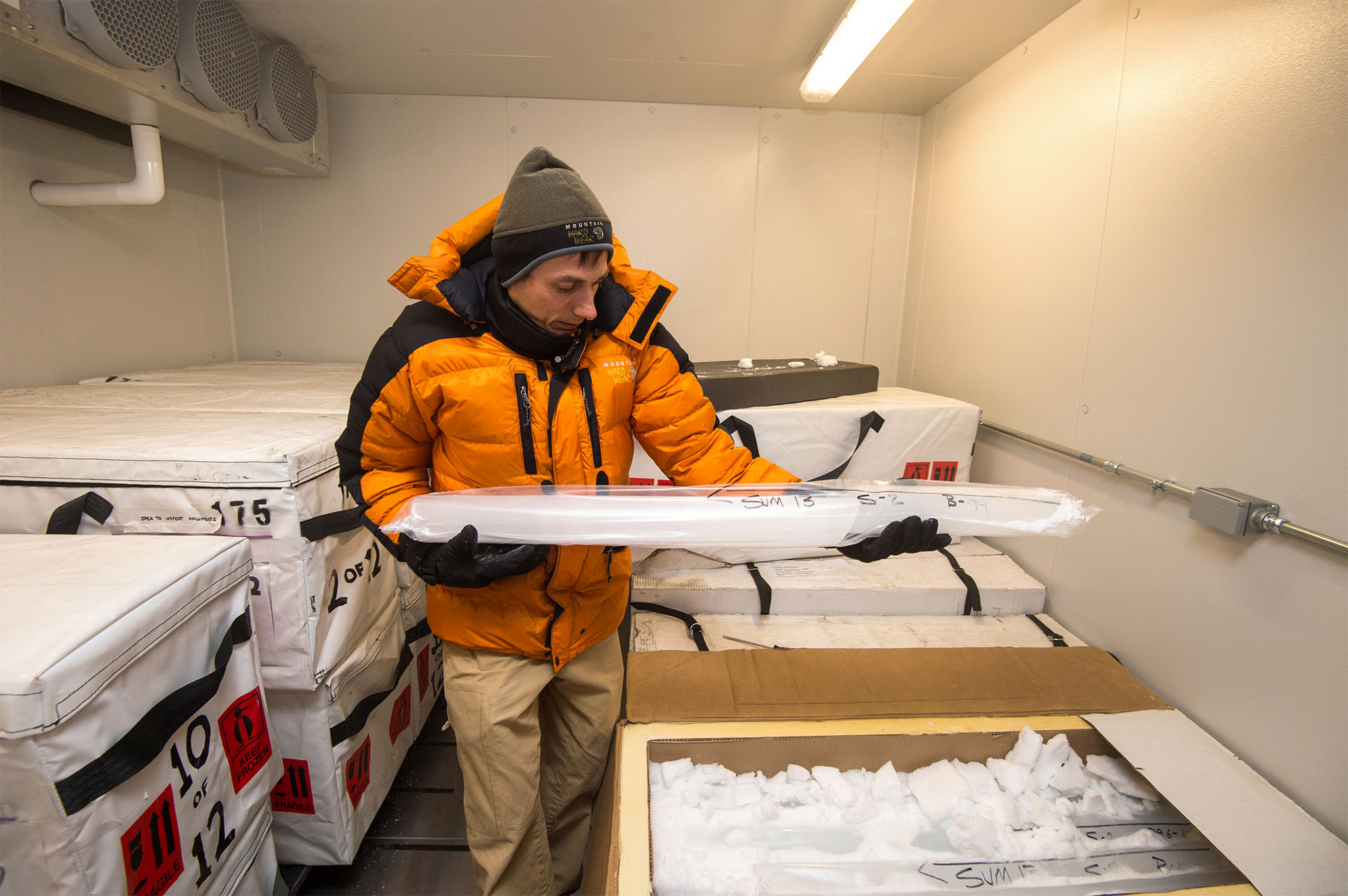 Vasilii Petrenko in an orange parka examines a long ice core above a collection of ice samples in an insulated laboratory.