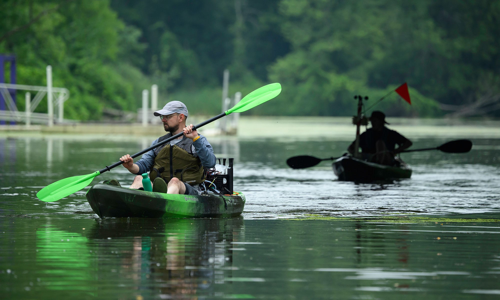 John Kessler paddles a green kayak equipped with scientific instruments on a calm waterway, with another kayaker behind him.