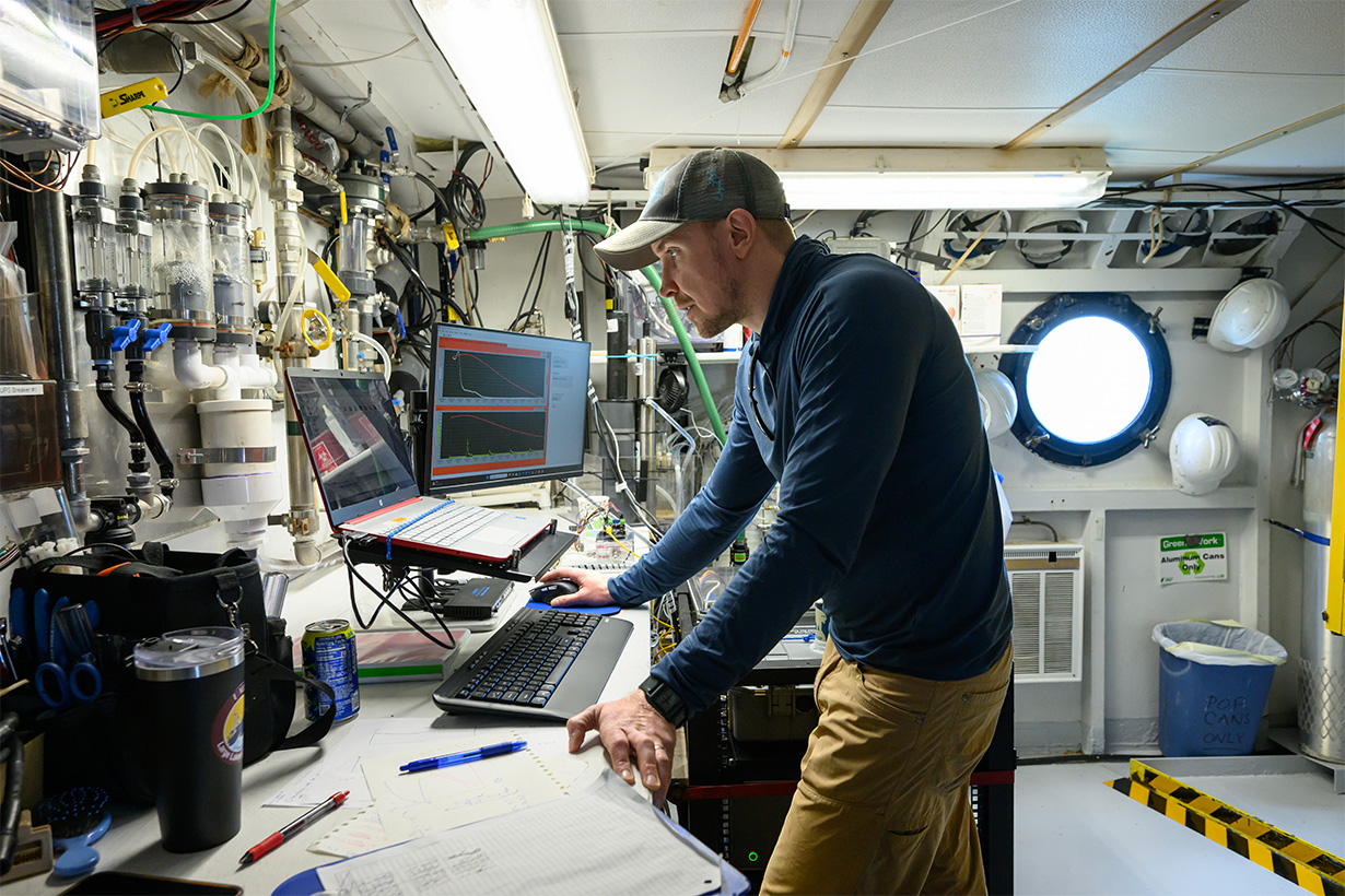 Thomas Weber works at a computer amid monitors, tubing, and scientific equipment aboard the R/V Blue Heron.