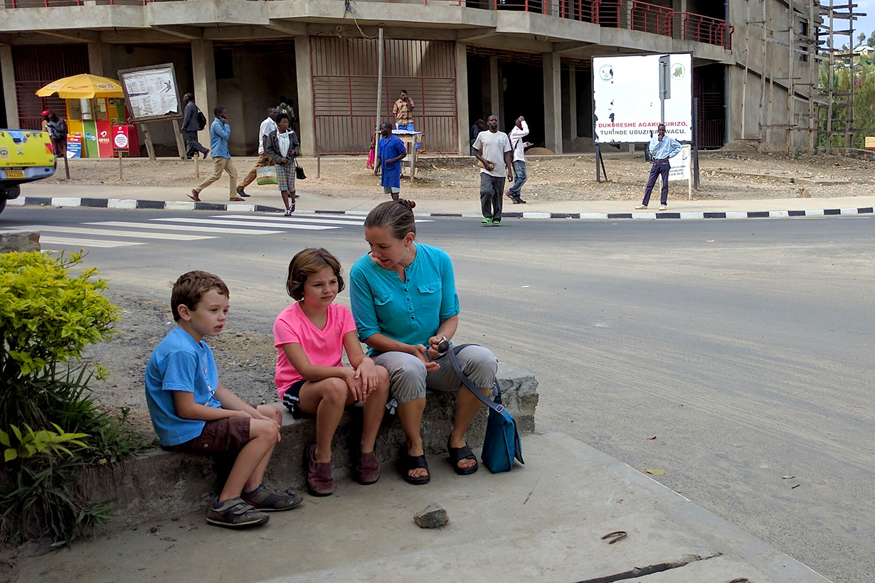Kristin Doughty and her two children sit next to a street in Rwanda lined with multistory buildings, with pedestrians and vehicles along the road.