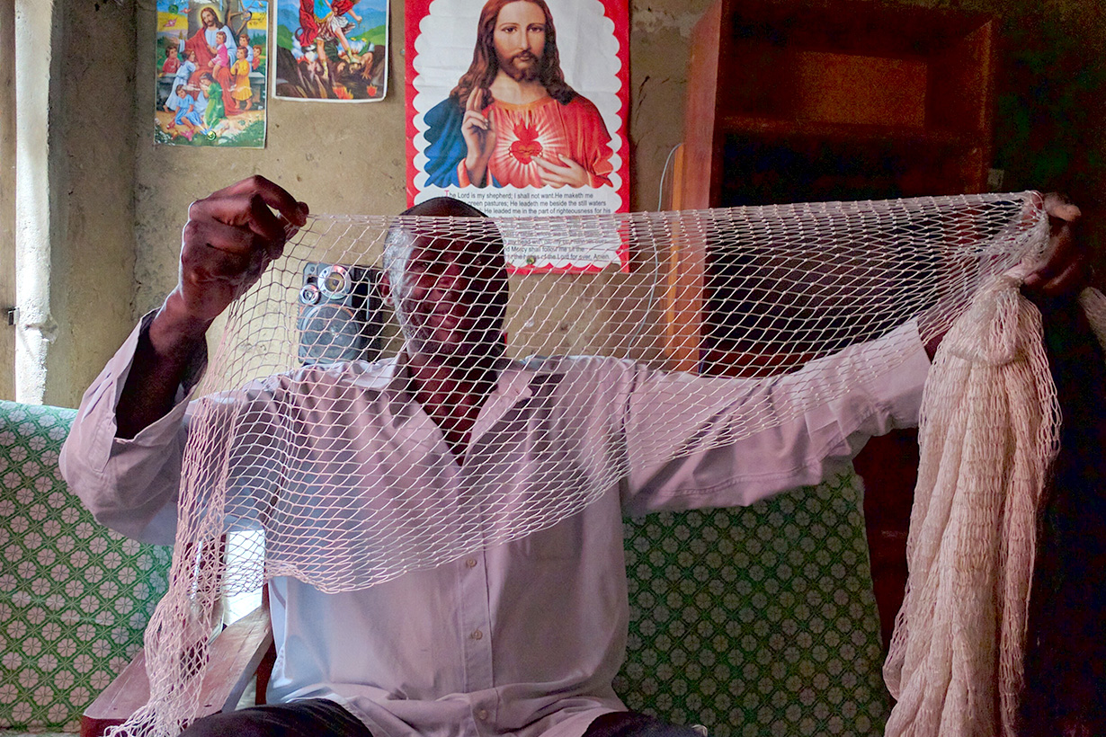 A person wearing a light-colored shirt is sitting on a patterned couch inside a rustic room while holding up a fishing net with both hands, with religious posters on the wall behind them.