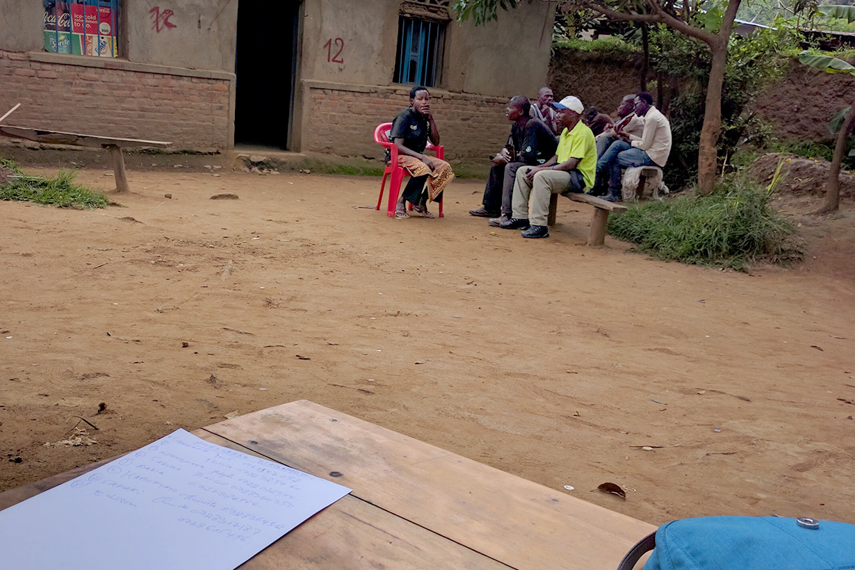 A group of people sit and talk outdoors near a building while a wooden table in the foreground holds a notebook, a pen, a sheet of paper, and a blue backpack.
