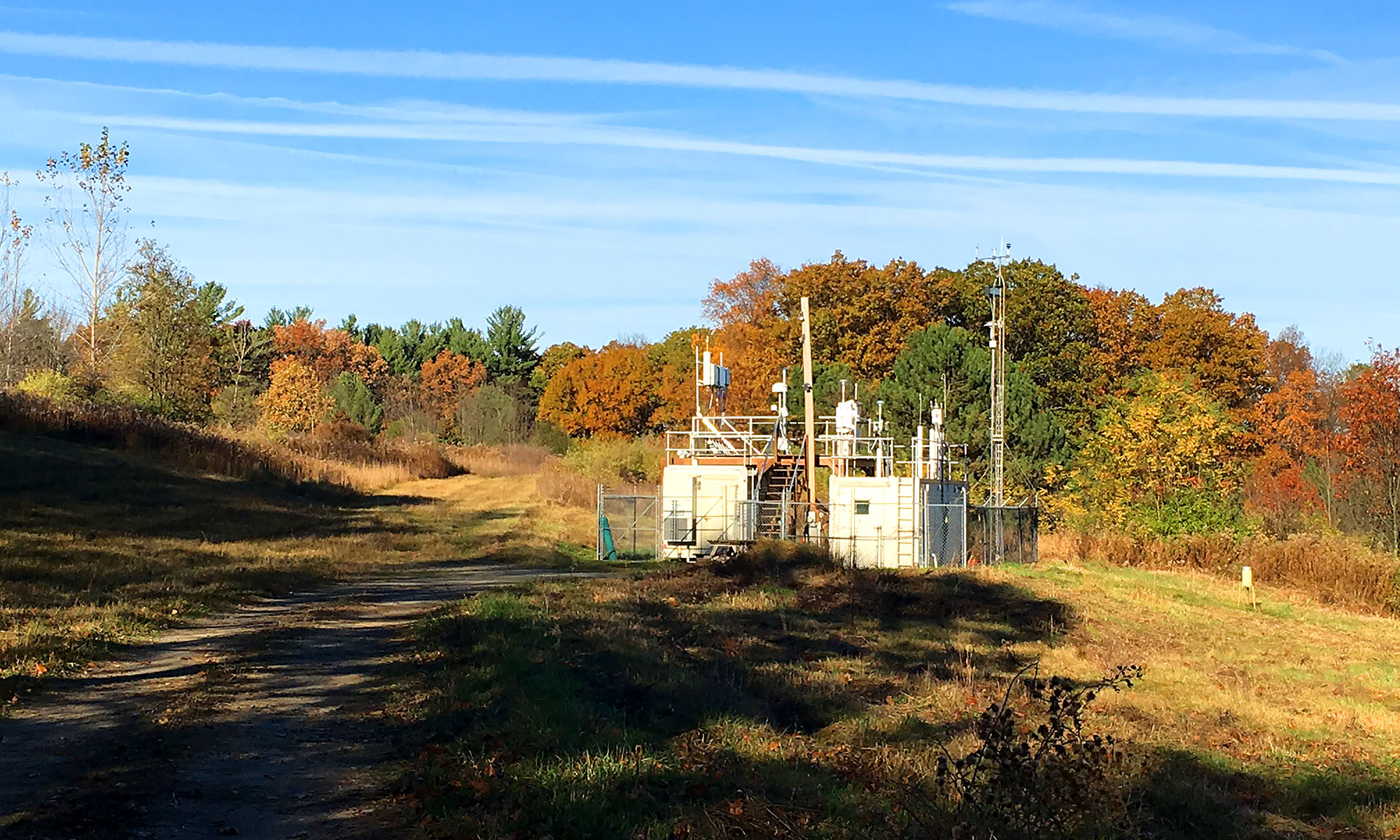A small greenhouse gas monitoring station stands beside a wooded landscape with colorful autumn foliage.