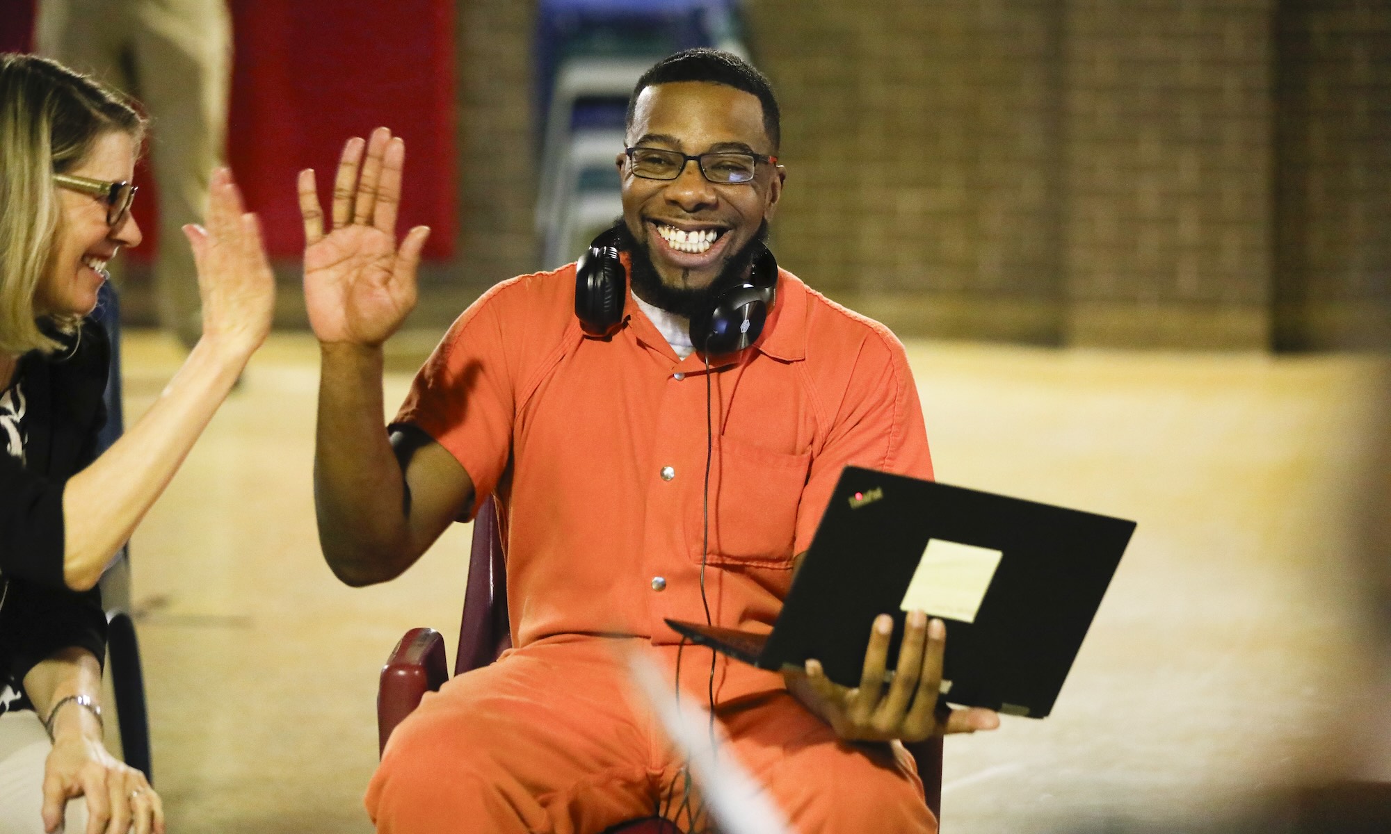 Inmate Micah smiles while sitting and holding a laptop and high-fiving a woman next to him.