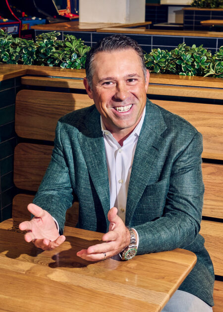 Shake Shack CEO Rob Lynch, wearing a green blazer, smiles and gestures with his hands while seated at a wooden restaurant table.