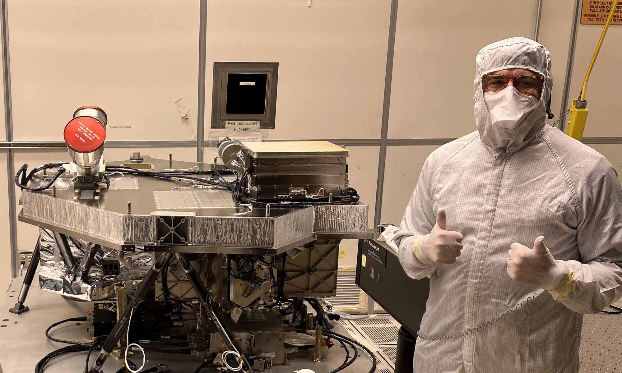Atmospheric scientist Sean Crowell gives two thumbs-up beside satellite equipment in a clean room, wearing a protective suit and mask.