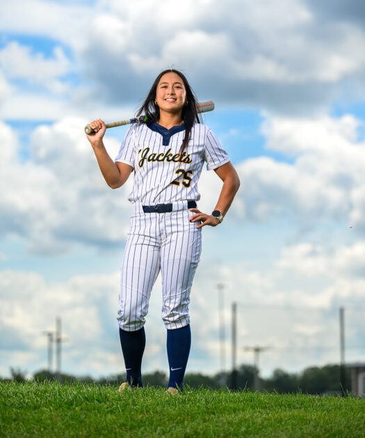 Riley Laygo dressed in a Yellowjackets softball uniform stands on grass with a baseball bat over her shoulder and blue sky with clouds behind her.